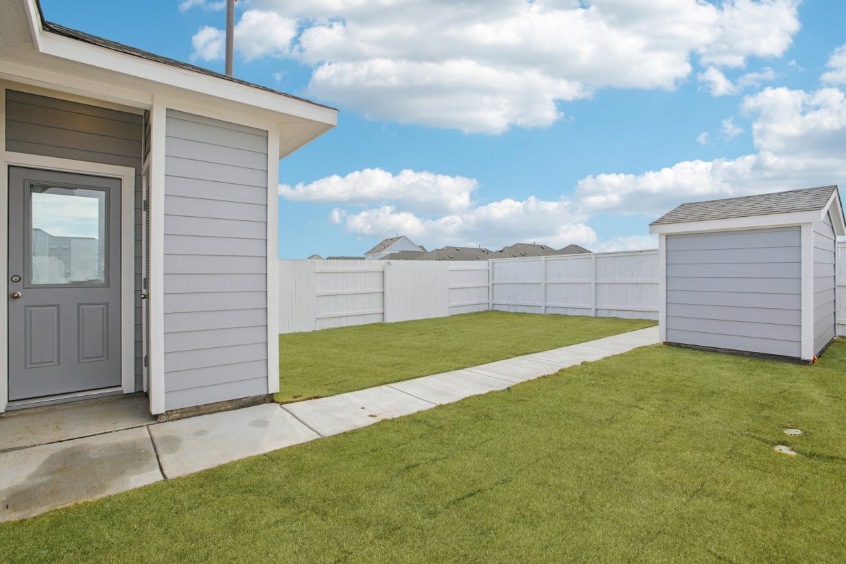 Image of a light grey house back exterior with a small grey shed, green grass, a white fence, and a blue sky in the background