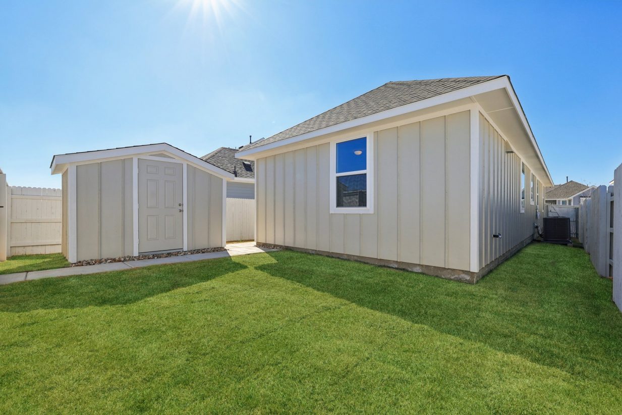 Image of the beige back exterior home with a detached shed, a green grass backyard and a blue sky in the background