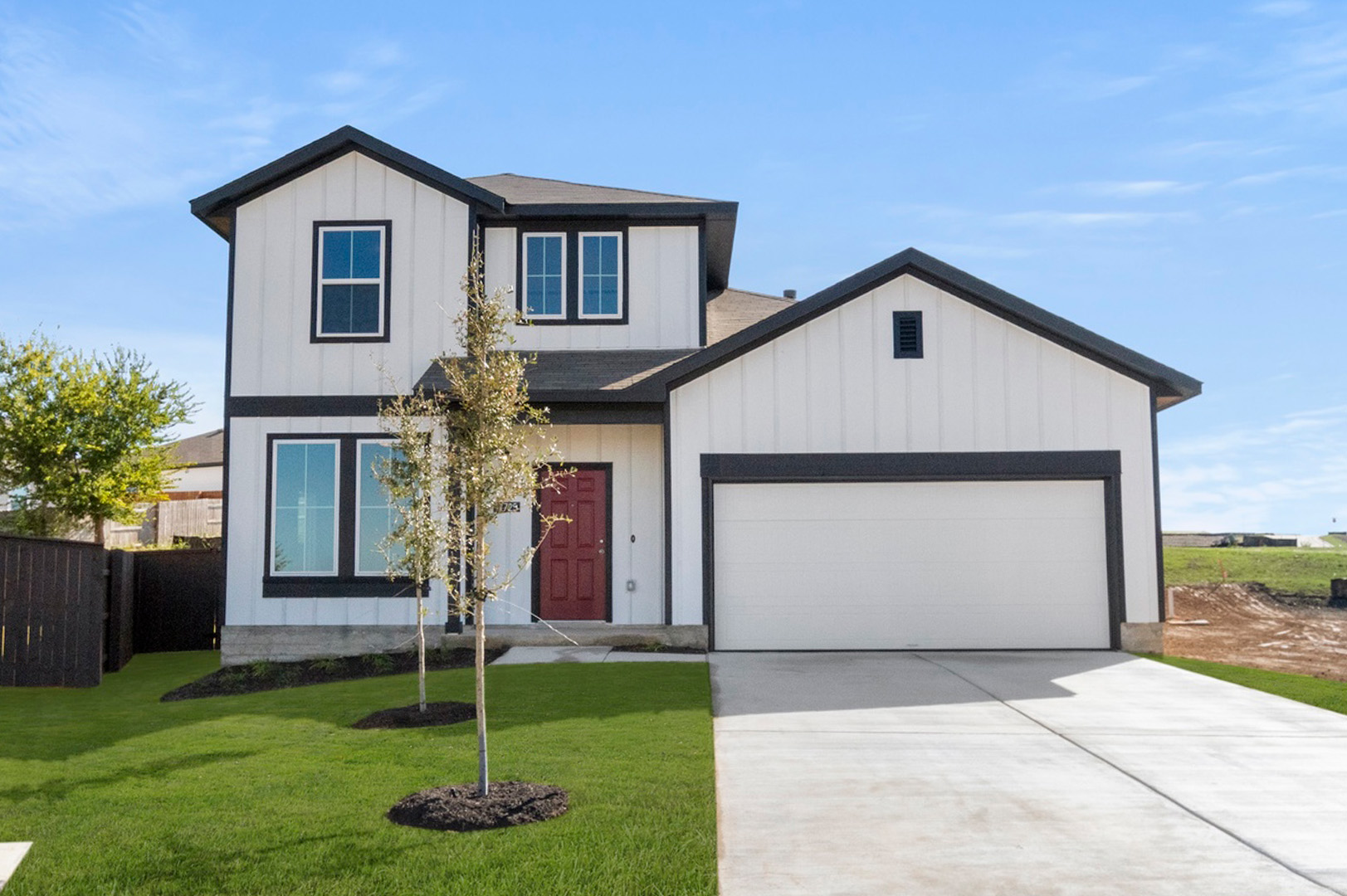 An image of the front of white farm-house styled two-story home with a red door, black framing, white garage door with green landscaping and a blue sky background.
