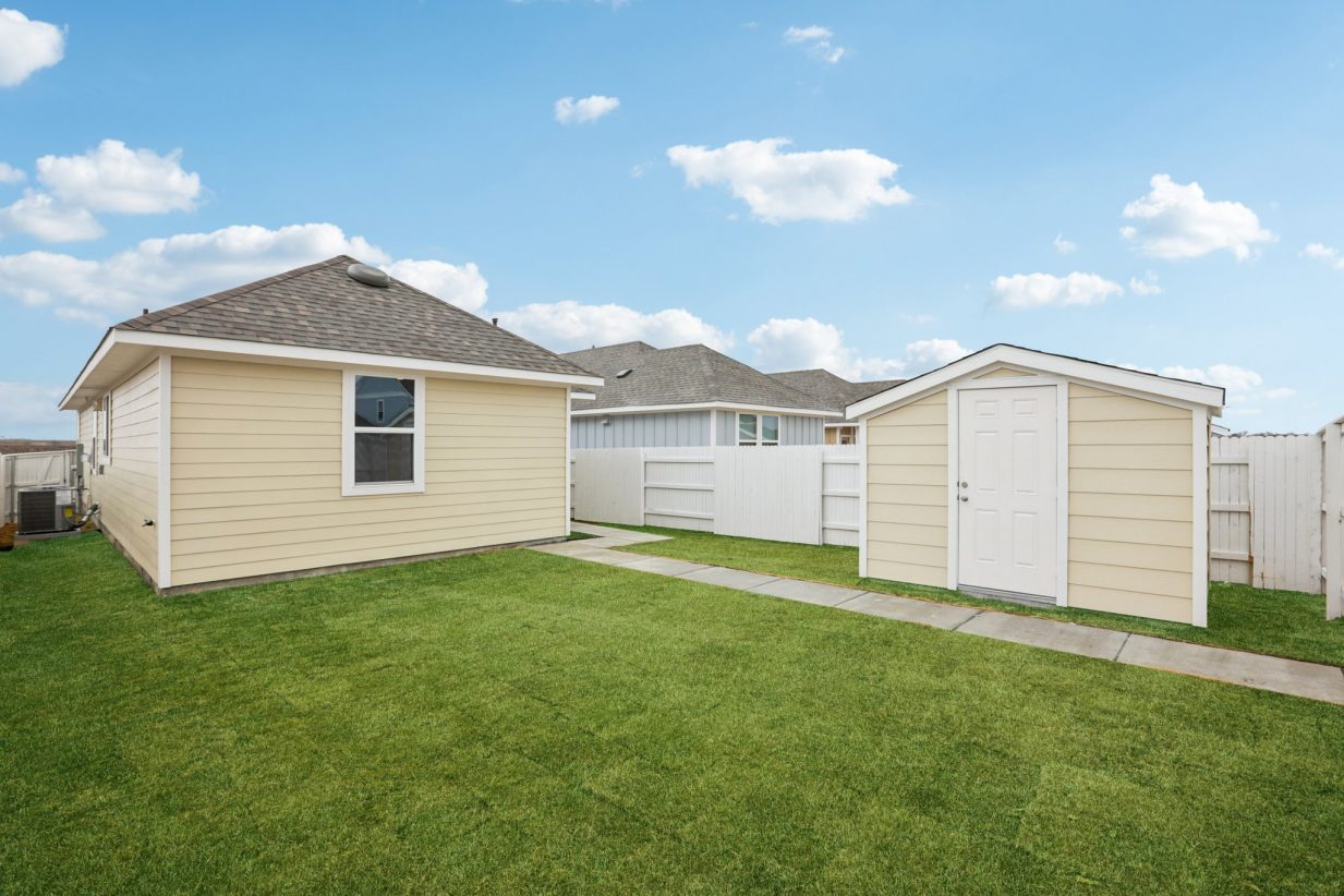 Image of a one story yellow house back exterior with a green grass backyard, a shed, a white wooden fence and a blue sky in the background