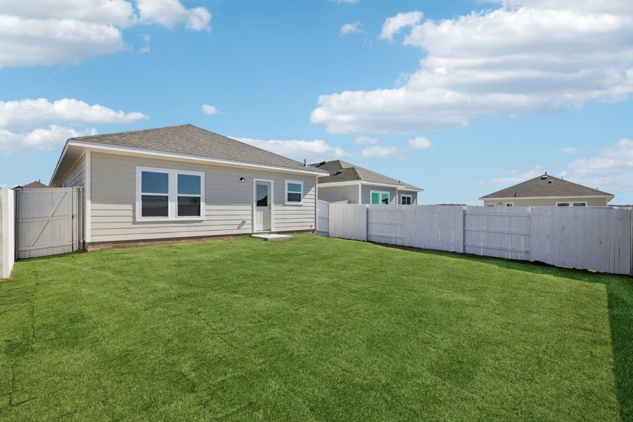 Image of a grey one story house back exterior with a green grass backyard, a white fence and a blue sky in the background
