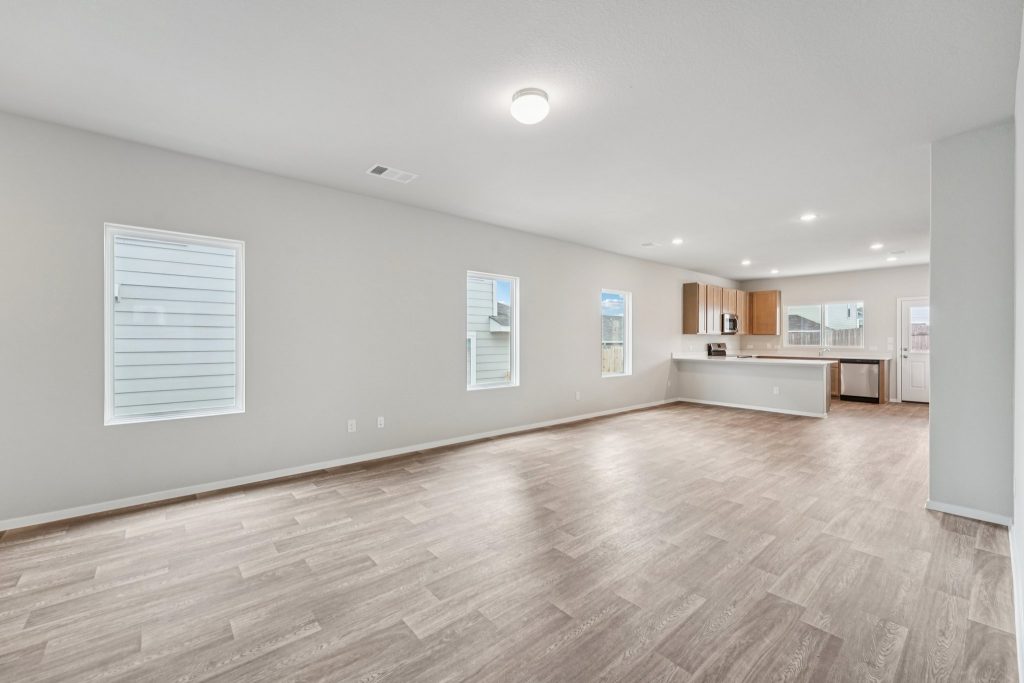 Image of a living area with vinyl flooring, light grey walls, windows and a a kitchen in the distance