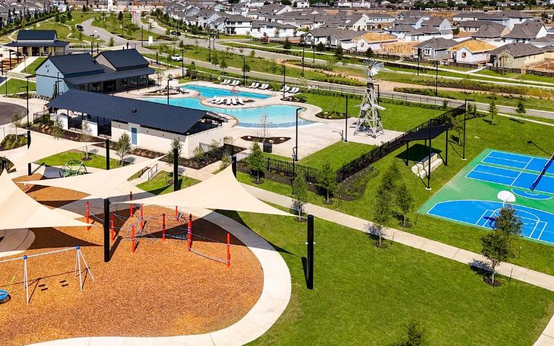 An aerial image of new home community with green landscape and a pool, pickleball court and playground with homes and the blue sky in the background