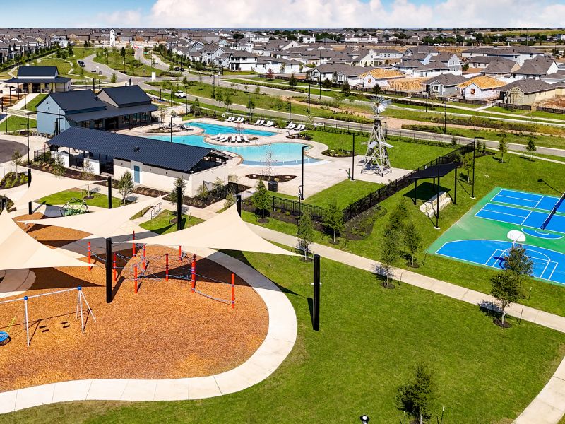 An aerial image of new home community with green landscape and a pool, pickleball court and playground with homes and the blue sky in the background
