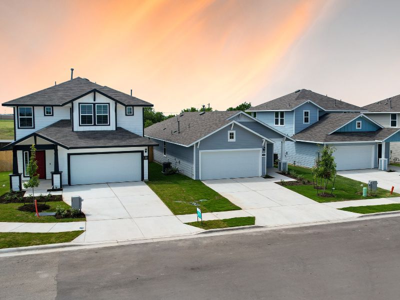 One-story and two-story new homes with a brown roof, concrete driveway, and green landscape with a yellow, blue, and orange sunset sky in the background.