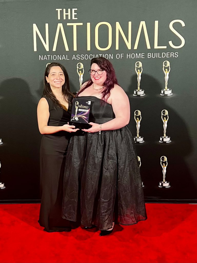 An image of two women on a red carpet. One with red hair and the other with brunette wearing black dresses in front of black backdrop with gold words "Nationals" and "National Association of Home Builders"