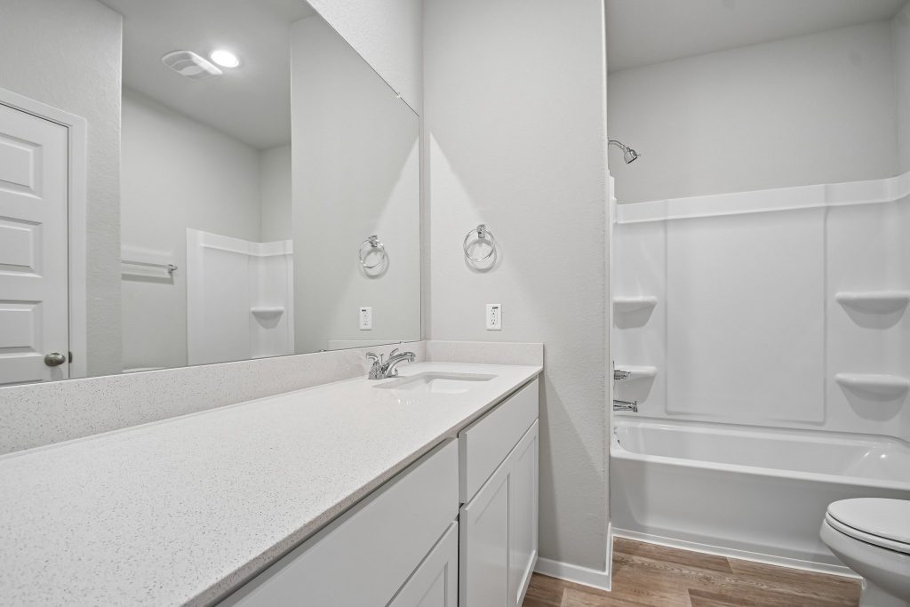 Image of home bathroom with chrome fixtures, white countertops, cabinets, bathtub and surround, toilet, and walls. Mirror across the vanity showing the door and towel bar over the toilet