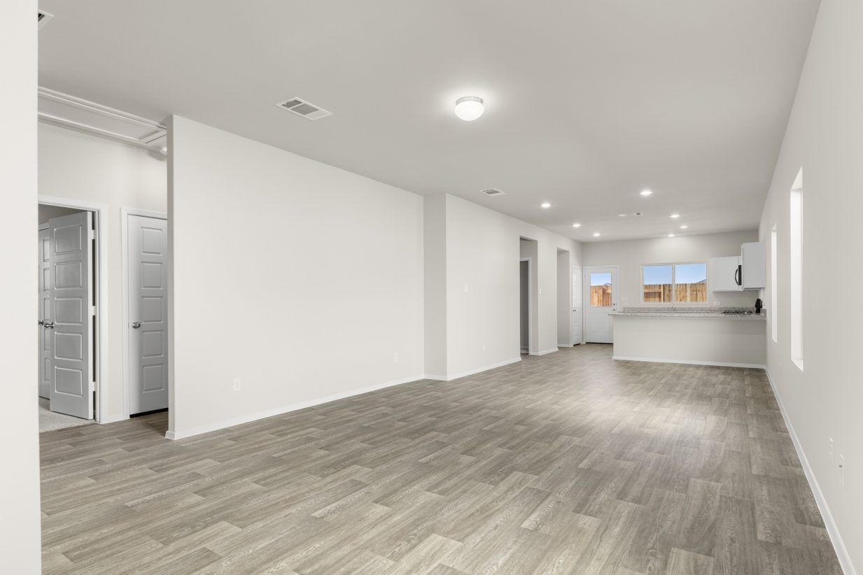 Image of home living spaces with wood-look flooring and light walls. Doorways and hallways in the left corner and kitchen in the back with multiple windows and door to the backyard