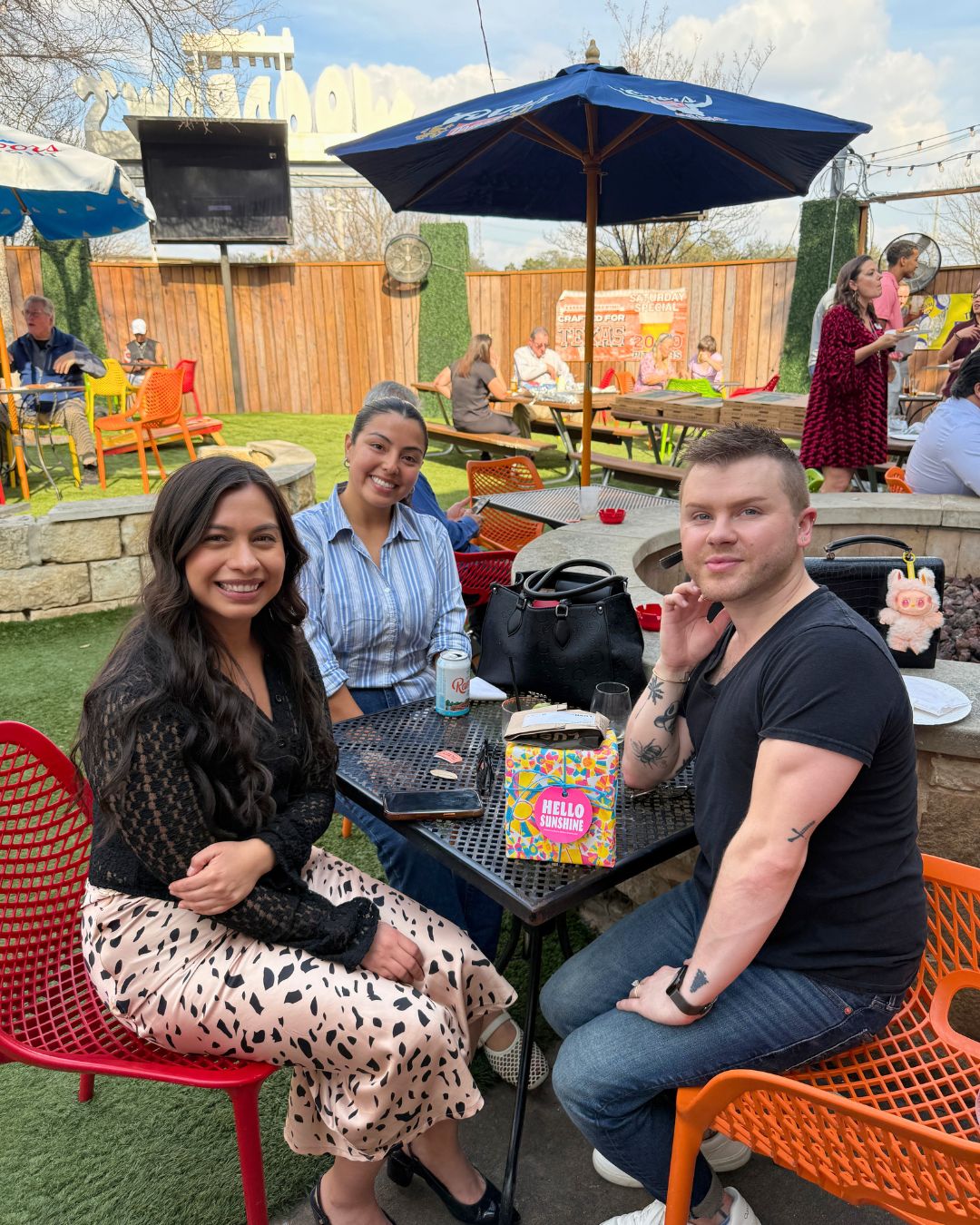 A picture of a group of people, two women and one man sitting down on orange chairs out side surrounded by other tables, green grass, and brown fence.