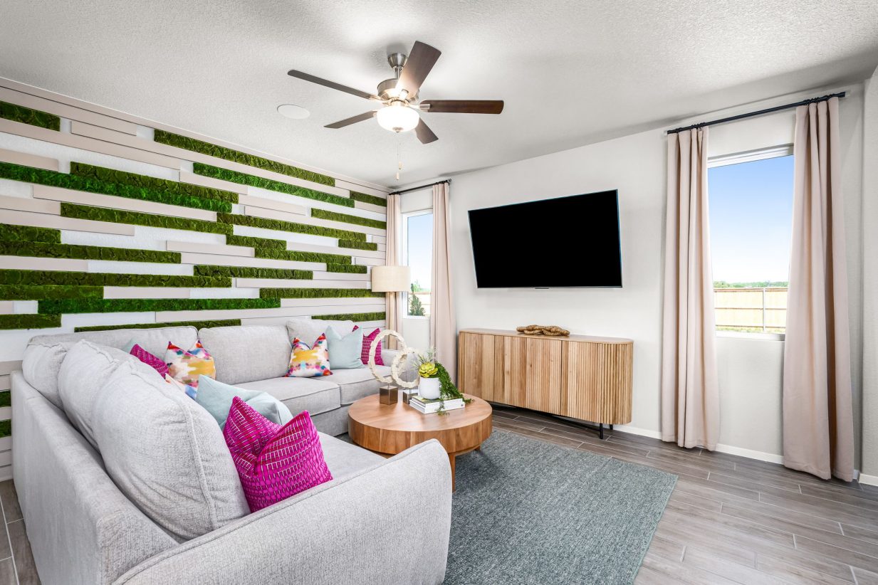 An image of living room with a white couch with bright pink pillows, tan coffee table, greenery, and a window with tan curtains.