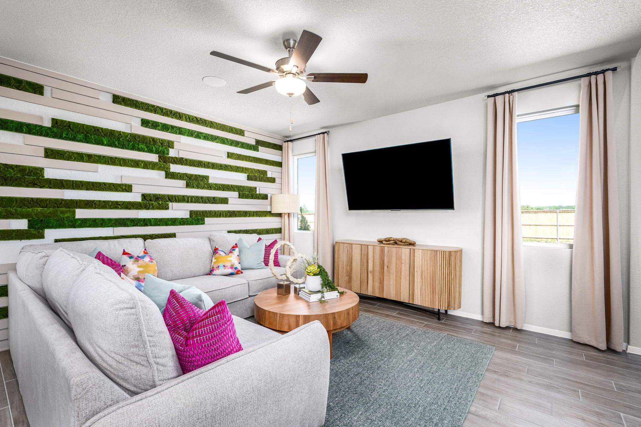 An image of living room with a white couch with bright pink pillows, tan coffee table, greenery, and a window with tan curtains.