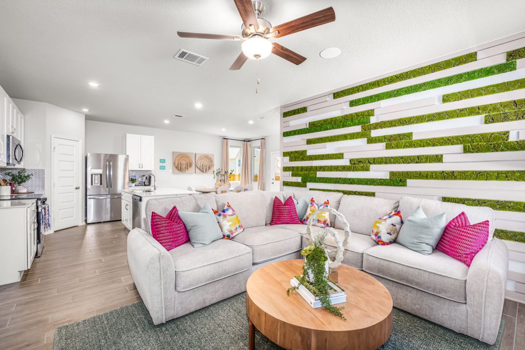 An image of living room with a white couch with bright pink pillows, tan coffee table, greenery, and the kitchen in the background with white counters and stainless steel appliances.