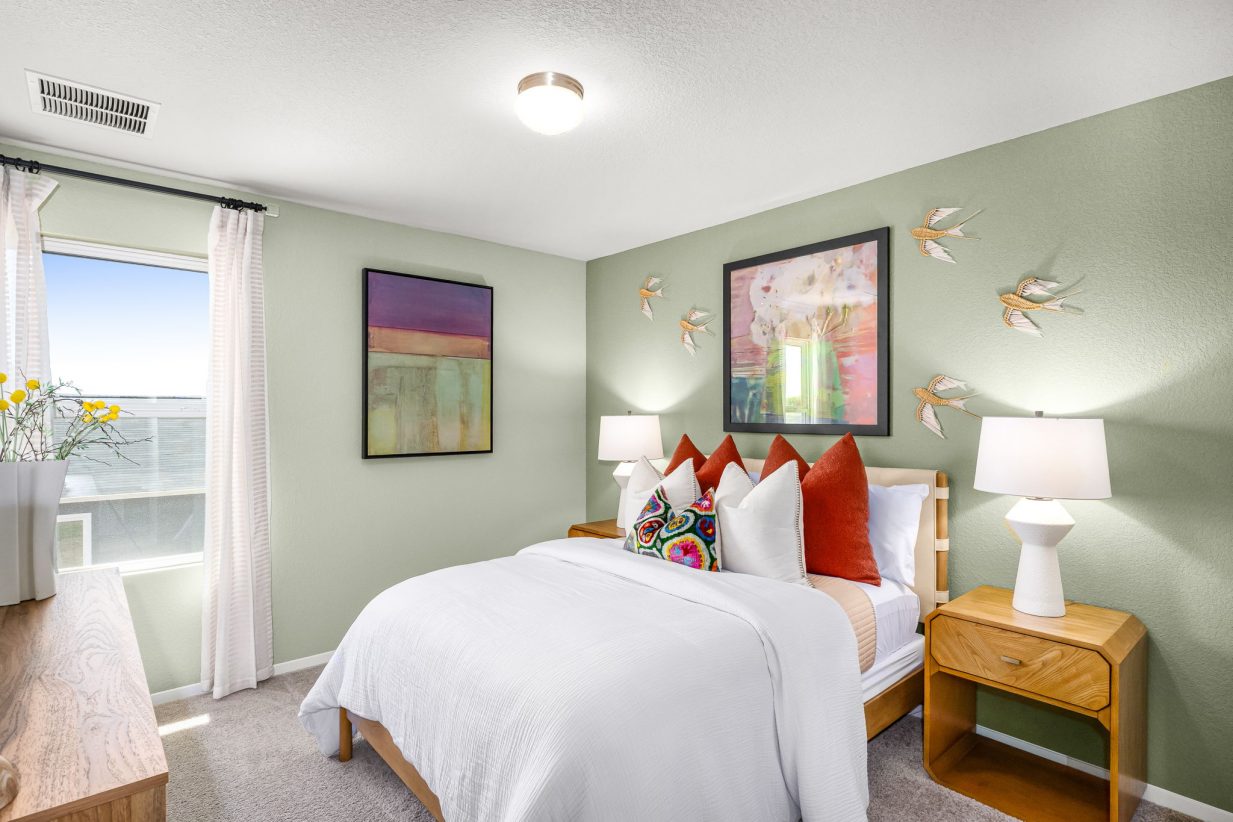an image of a bedroom with green walls, colorful art on the wall, brown side table, white bed with colorful pillows, and a window.