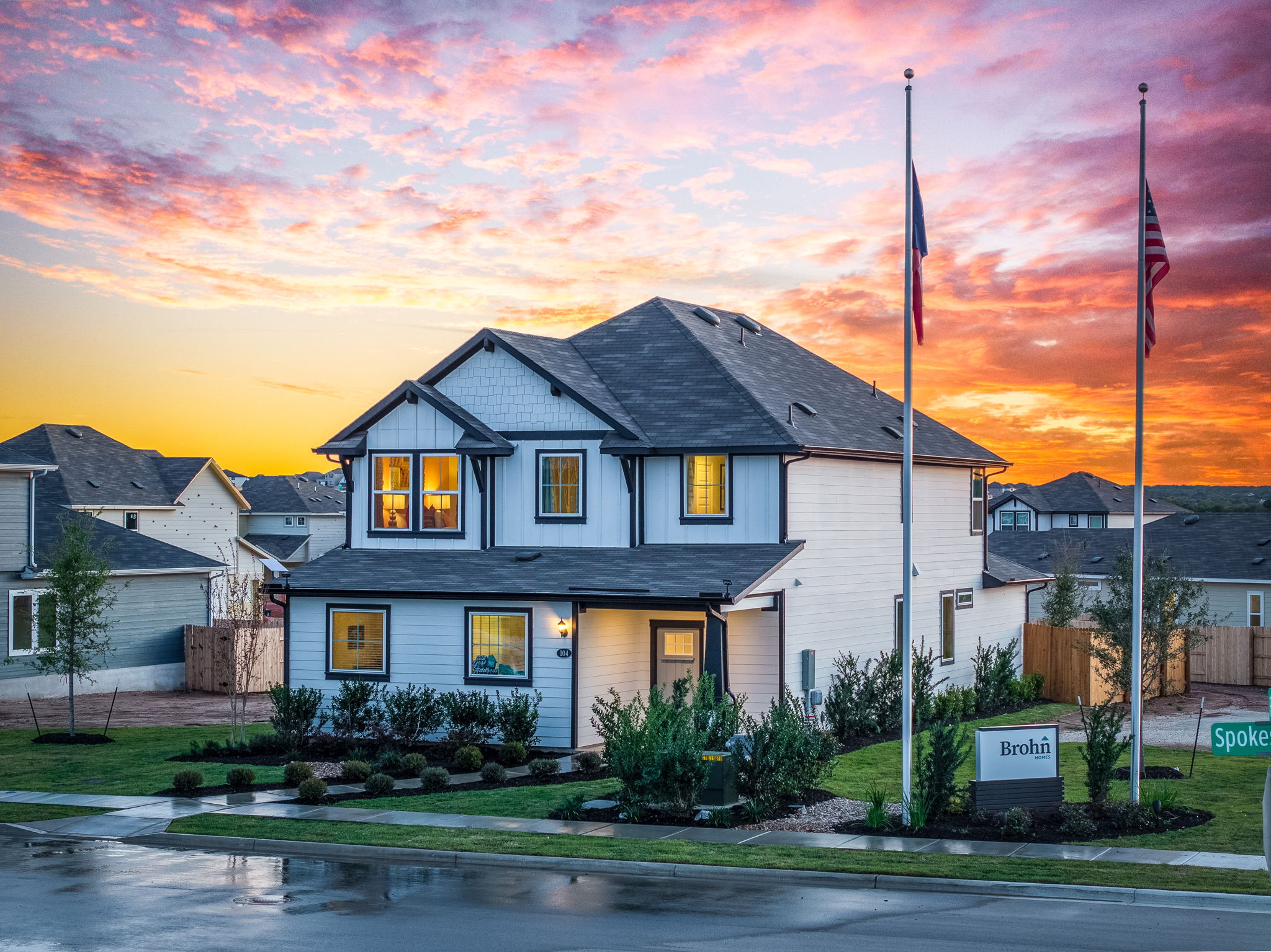 An image of two story white house with black trimming and a brown roof surrounded by green landscape with bushes with a sun setting background with pink, orange, yellow, and blue.