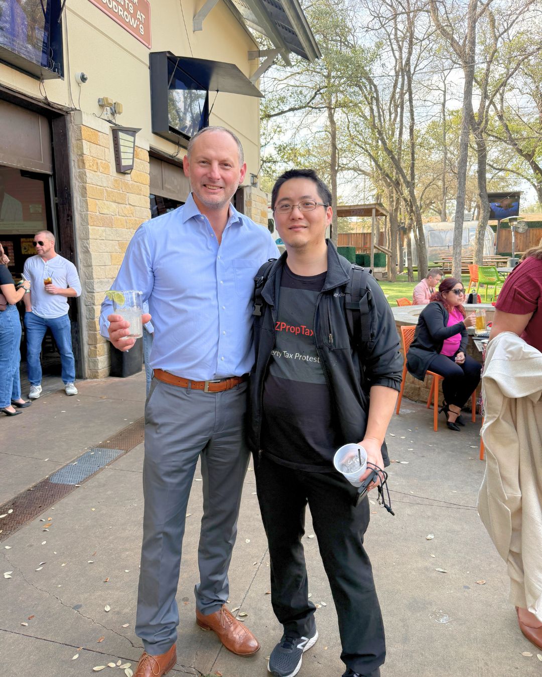 A picture of two men holding drinks and smiling outside with concrete floor and building and trees in the background.