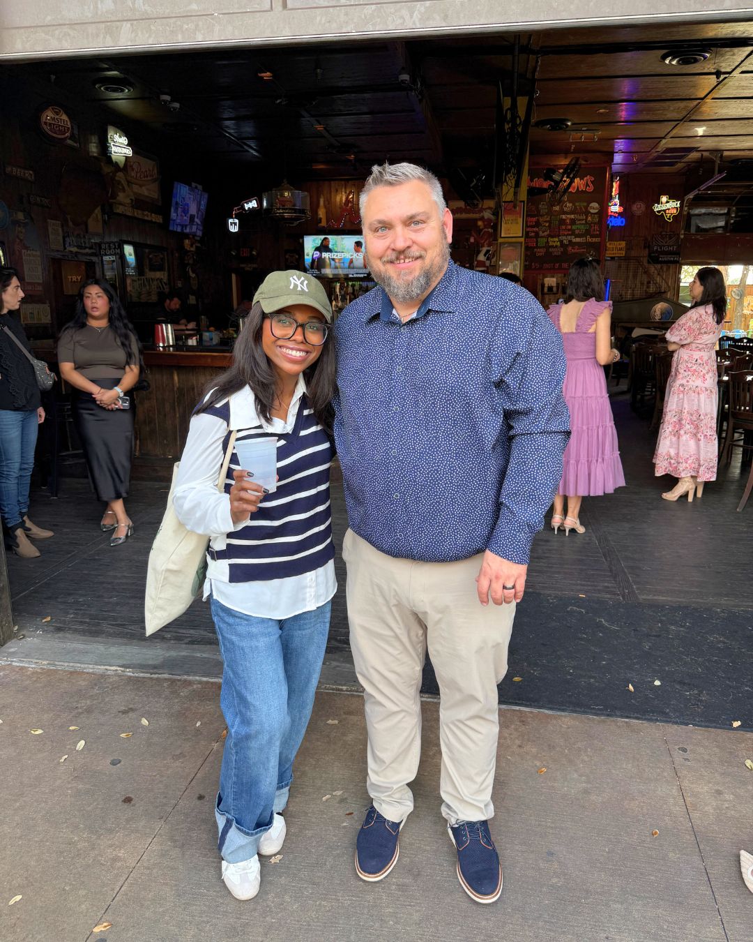 A picture of a woman and a man wearing casual clothes and side hugging on concrete with people and a bar in the background.