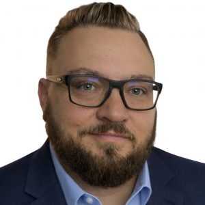 Headshot of man with light smile wearing black glasses, blue shirt, and dark blue jacket