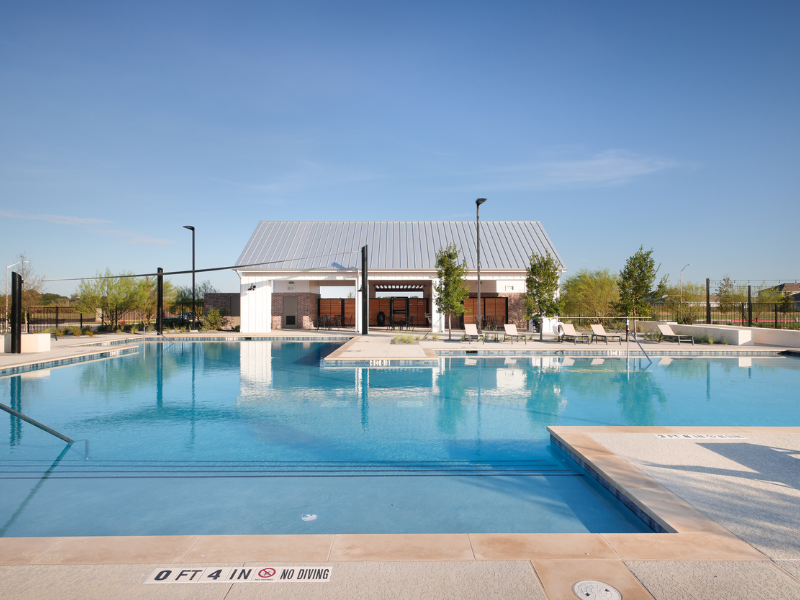 An image of pool, surrounded by concrete foundation with some green plants with a white house structure and a blue sky behind it.