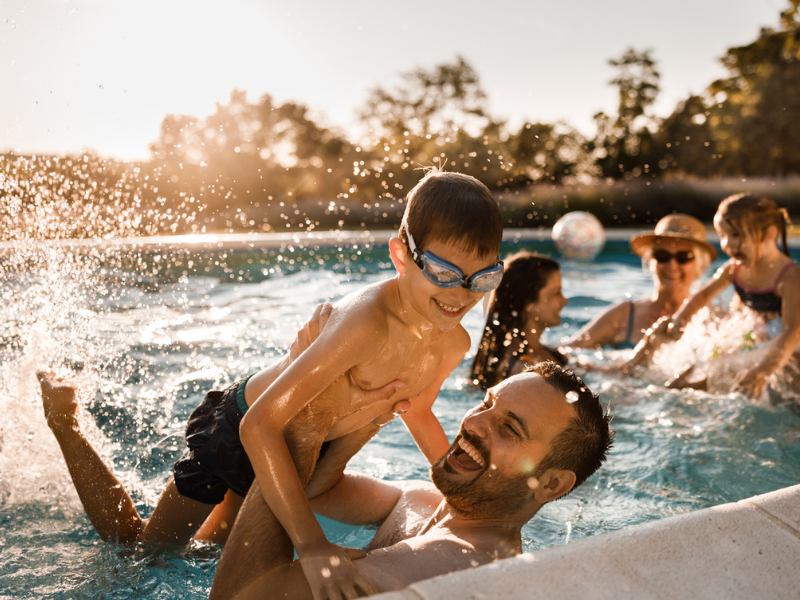 Father and son playing in the swimming pool with other people in the background.
