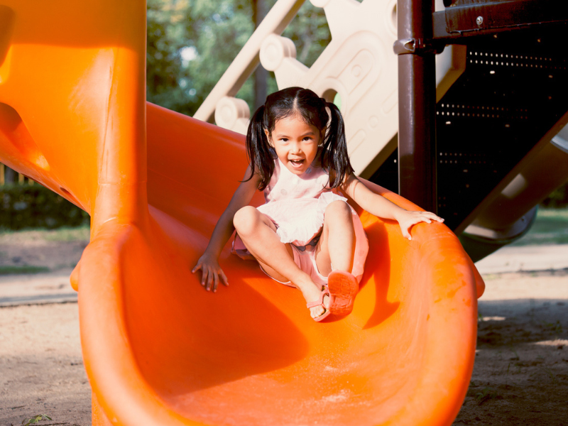 Asian little girl in a pink dress sliding down an orange slide.