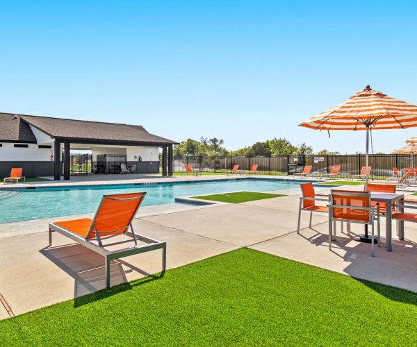 Image of community pool with orange chairs and orange and white umbrellas with amenity center in the back.