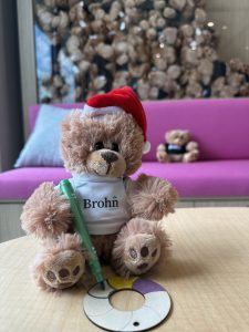 Image of teddy bear in white Brohn Homes shirt and Santa hat holding a green marker coloring a lifesaver ornament. Bear sitting on pink couch in front of a bear display