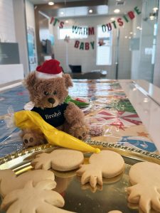 Image of teddy bear in black Brohn Homes shirt and Santa hat holding a bag of yellow icing decorating pineapple shaped cookies. Banner in background reads it's warm outside baby