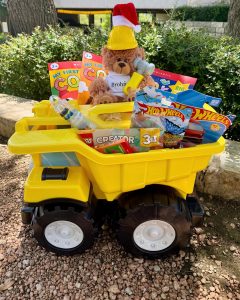 Image of teddy bear wearing white Brohn Homes shirt, yellow hard hat, and santa hat sitting in a toy dump truck filled with toys