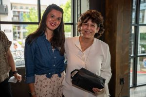 Image of two woman standing together and smiling. One woman is holding a black, small bag