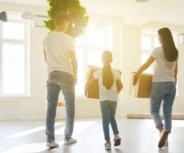 A Family with woman, man, and little girl wearing white shirts and blue jeans carrying boxes inside a home with many windows letting in sunshine.