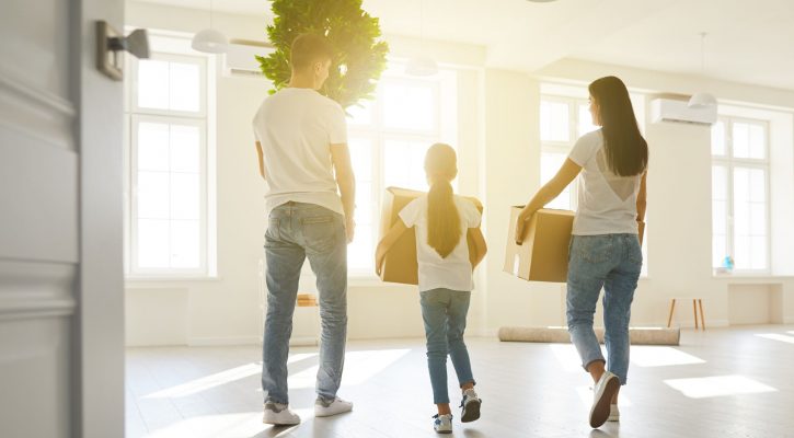 A Family with woman, man, and little girl wearing white shirts and blue jeans carrying boxes inside a home with many windows letting in sunshine.