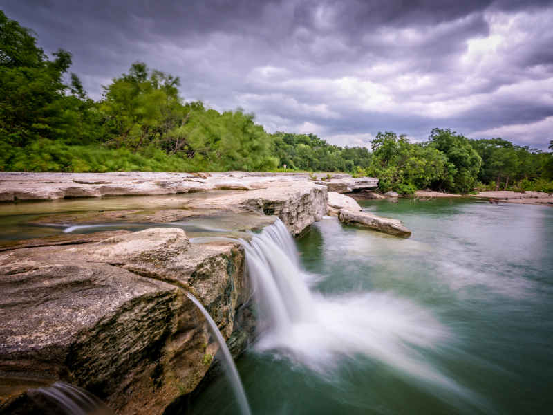 An image of a beautiful water fall running over to the river below with a dark cloudy sky in the background.