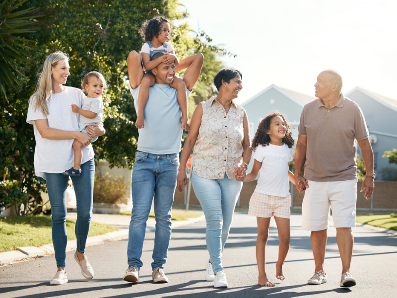 A mixed family with a mom, dad, two children, and grandparents walking on the concrete street with greenery, trees, and house figures in the background.
