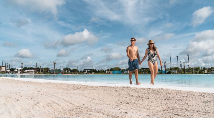 An image of couple wearing bathing suits and sunglasses walking down a beach in the blue water with a blue sky with clouds in the background.