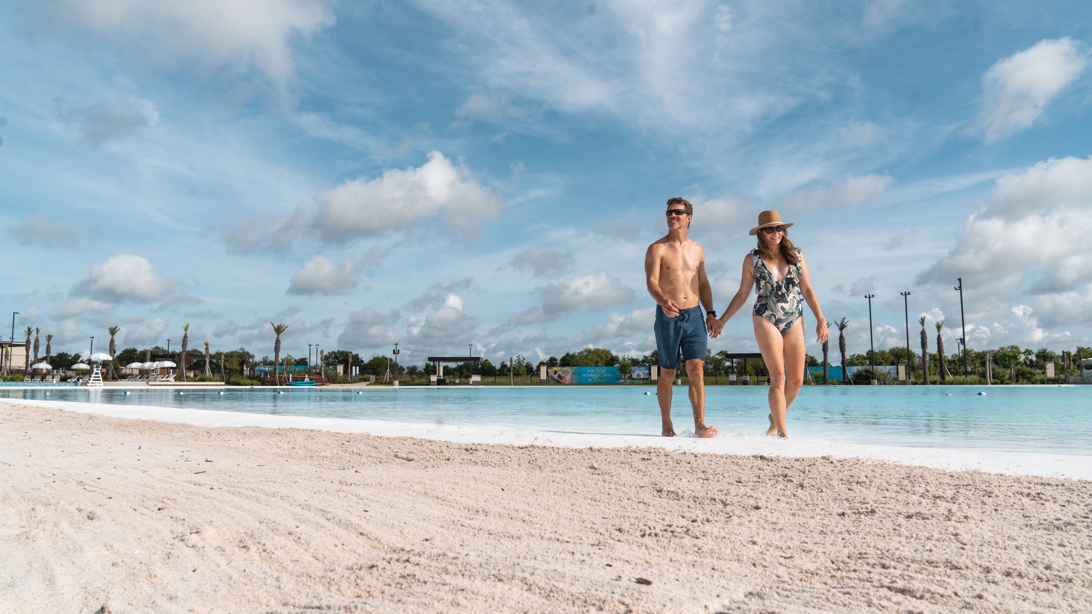 An image of couple wearing bathing suits and sunglasses walking down a beach in the blue water with a blue sky with clouds in the background.