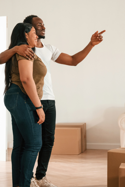 A african american man with a white t-shirt arm wrapped around african american woman with a army green shirt with hard wood floor and boxes in the background.
