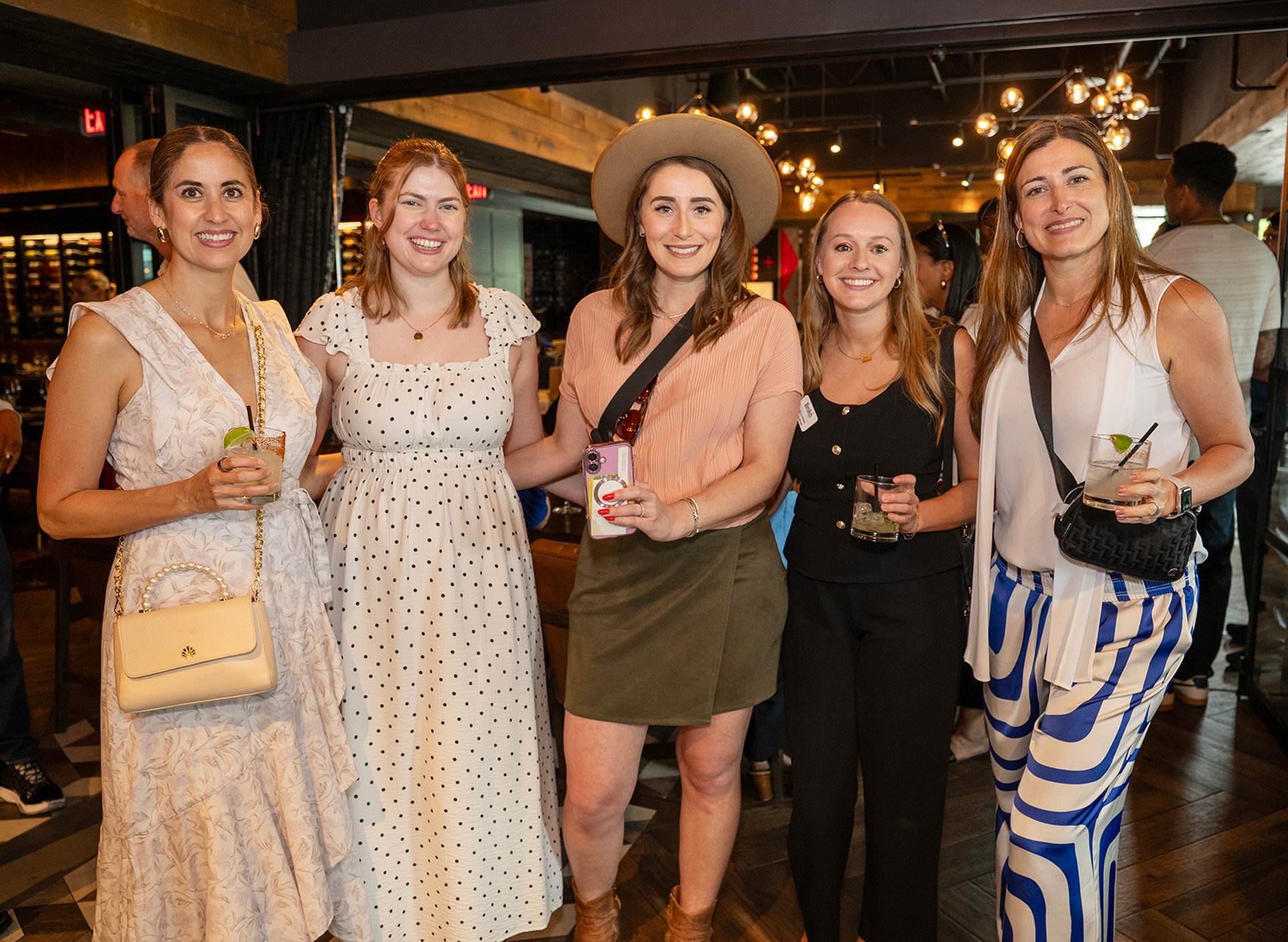 A group of women smiling and huddled together with drinks in a bar like setting with lights and wood paneling in the background.