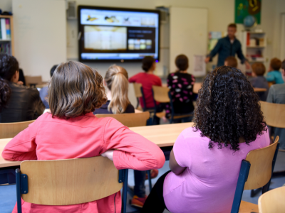 An image of male and female elementrary students sitting down at tan desks with their heads forward facing a screen and the male teacher.