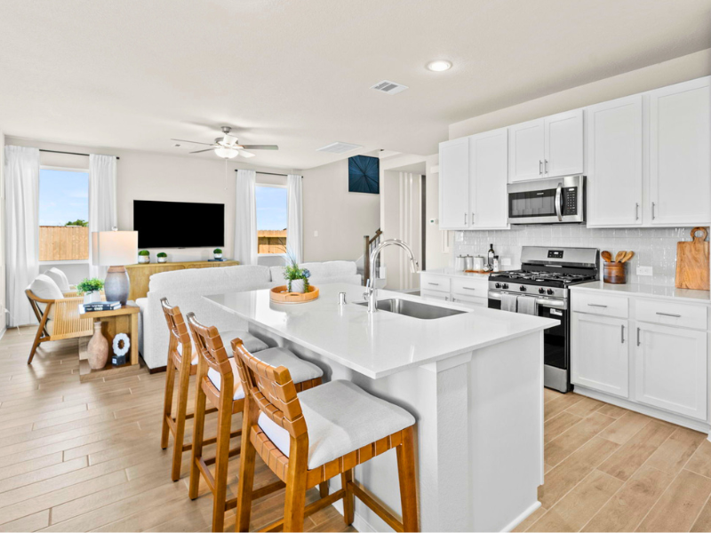 An interior image with a white counters, a white island, white cabinets, stainless steel appliances, with a living room area in the background with a black TV, white couch, ceiling fan, and windows.
