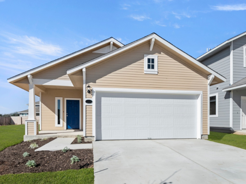 Image of the front exterior of a yellow one-story house with a concrete drive way and green grass with landscaping with a blue sky in the background.