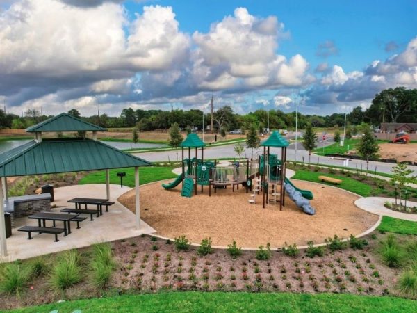 An aerial image of a green playscape with slides,bridge, a pavilion with benches and picnic tables, greenery, and a background with a blue sky with clouds.