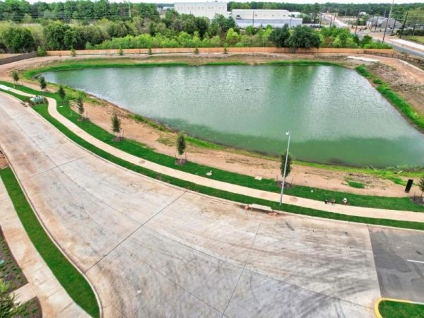 An Aerial image of blue and green pond surrounded by trees, greenery, and dirt, next to a concrete paved road.