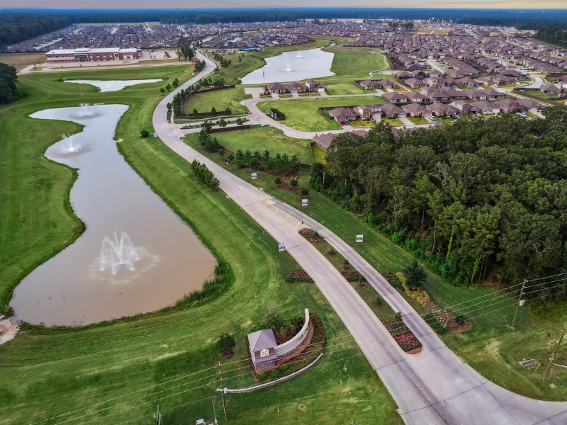 An aerial view of neighborhood with a brown sign, big pond surrounded by grass, trees, and a road that leads to homes in the background.