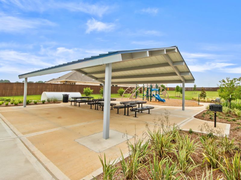 An image of pavilion with a metal roof, concrete patio, black picnic table and bench, and a blue sky with white clouds.