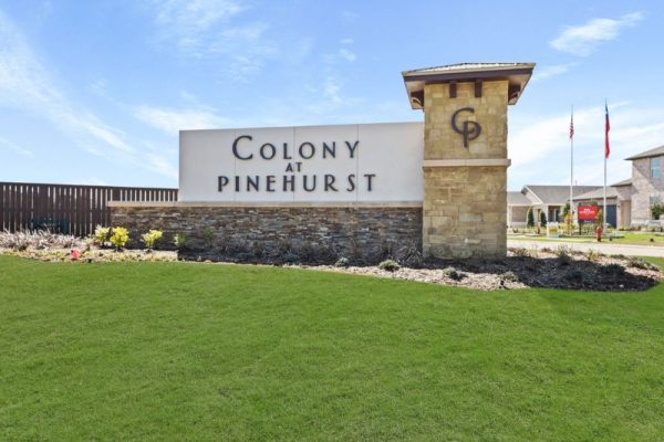 An exterior image of a white sign with brown text that says "Colony at Pinehurst," set against a brick-stone basin, green grass, pebble landscaping, and a brown fence, with a blue sky and white clouds background.