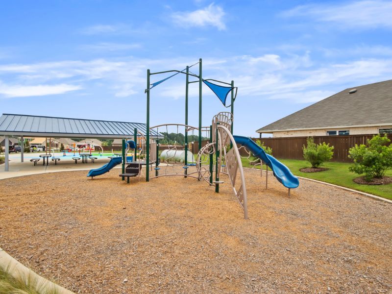 An image of blue an tan playscape with a blue slide, wood chips, with a pavilion with a metal roof and a blue sky with white clouds in the background.