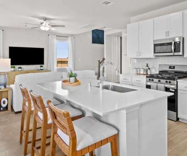 Image of a kitchen looking into a living room with white counters, a stainless-steel oven and built-in microwave, an island, a white couch, a black TV, wood-look vinyl flooring, windows, and white walls.