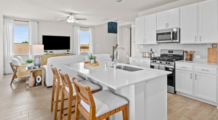 Image of a kitchen looking into a living room with white counters, a stainless-steel oven and built-in microwave, an island, a white couch, a black TV, wood-look vinyl flooring, windows, and white walls.