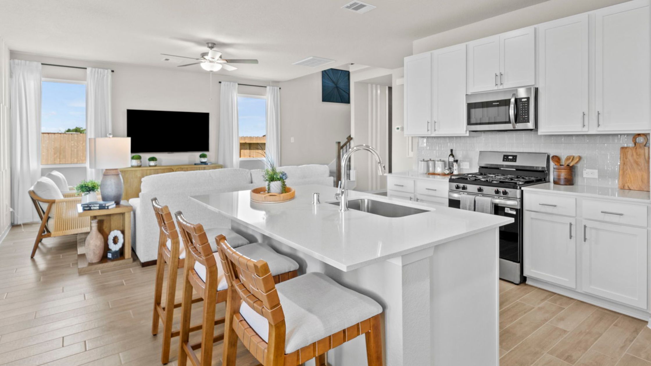 Image of a kitchen looking into a living room with white counters, a stainless-steel oven and built-in microwave, an island, a white couch, a black TV, wood-look vinyl flooring, windows, and white walls.
