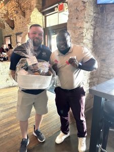 Photo of two men standing and smiling. The man on the left is holding a white gift basket